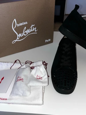 Svarta Louboutin sneakers med nitar - Säljer ett par svarta sneakers från Christian Louboutin i mocka med svarta nitar på tån och den klassiska röda sulan. Skorna har svarta skosnören och en låg silhuett som ger en clean och edgy look. Perfekta för dig som vill sticka ut med exklusiva detaljer.