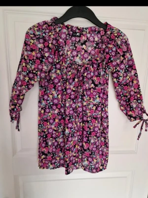 Blommig blus från JC med trekvartsärm - Säljer en färgglad blommig blus från JC med trekvartsärmar och knytband vid ärmslut. Blusen har en lila bas med blommönster i rosa, vitt, gult och blått. Materialet är lätt och luftigt, perfekt för att skapa en avslappnad och trendig look. Går att justera uringningen med hjälp av snörning