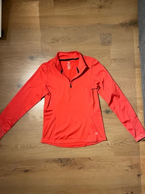 Röd långärmad träningströja SOC - Snygg röd långärmad träningströja från SOC med halv dragkedja framtill och hög krage. Tillverkad i ett lätt och stretchigt funktionsmaterial som passar perfekt för löpning eller gym. Diskret logga på bröstet och svart detalj vid dragkedjan.