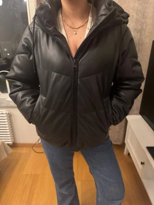 Svart pufferjacka med huva - Snygg svart pufferjacka med dragkedja och huva. Jackan har en stilren look med quiltade partier och två fickor framtill. Perfekt för kalla dagar och enkel att matcha med olika outfits. Jackan har en något boxig passform och är vadderad för extra värme.