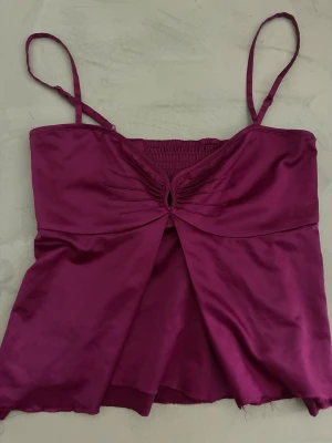 Lila satinlinne med smala axelband - Snyggt lila linne i glansigt satinmaterial med smala justerbara axelband och rynkdetalj framtill. Linnet har en liten öppning vid bysten och smock i ryggen för skön passform. Perfekt för att lyfta vilken outfit som helst.