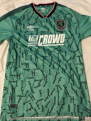 Grön FilthyFellas tröja från Umbro - Säljer en grön sporttröja från Umbro med FilthyFellas-logga, skapad av den kända UK podden med samma namn. Tröjan har svarta och rosa detaljer vid kragen och sponsortryck 'THE CROWD' på framsidan. Perfekt för fotboll eller streetwear.