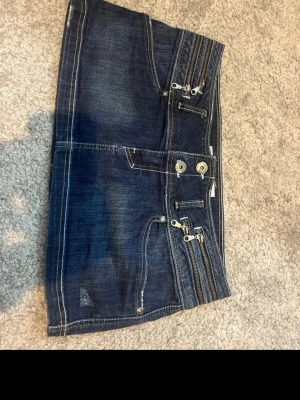 Mörkblå jeanskjol med dragkedjor - Säljer en mörkblå jeanskjol från Trendy Miss med flera dekorativa dragkedjor och knappar framtill. Kjolen har kontrastsömmar och klassisk femficksdesign. Perfekt för dig som gillar en edgy och cool stil.