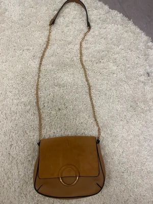 Brun axelväska med guldkedja - Snygg axelväska i brunt läder med guldkedja och justerbar axelrem. Väskan har en rund detalj på framsidan och lock i mocka-look. 