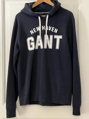 Mörkblå hoodie från GANT - Mörkblå hoodie från GANT med vit text 'NEW HAVEN GANT' på bröstet. Klassisk modell med huva och vita dragsnören. Tillverkad i mjukt bomullsmaterial och har långa ärmar samt ribbade muddar. Perfekt för en avslappnad och stilren look.  Kommer från fräscht djur ock rökfritt hem.