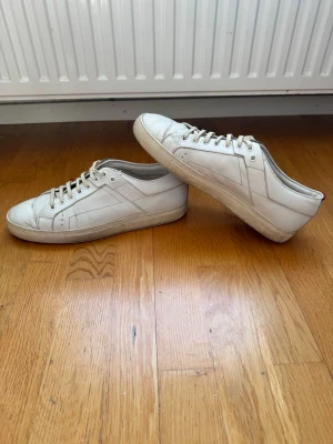 Vita sneakers från Hugo Boss - Säljer ett par stilrena vita sneakers från Hugo Boss. Skorna har klassisk låg profil, vita snören och diskreta sömdetaljer. Tillverkade i skinn med röd detalj bak på hälen och logga på insidan. Perfekta för en clean och enkel look.