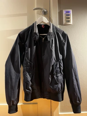 J.Lindeberg vårjacka - Stilren mörkblå vindjacka med dragkedja framtill och två stora fickor med knappar. Jackan har ribbade muddar vid ärmslut och midja samt en sportig krage med vita ränder. Perfekt för blåsiga dagar och enkel att matcha till olika outfits.