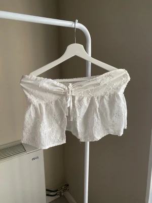 Vit offshoulder blus med spetsdetaljer - Supersöt vit offshoulder blus med broderade blommor och spetsdetaljer. Toppen har en kort, croppad passform och knyt framtill för extra stilpoäng. Perfekt för varma dagar och ger en romantisk vibe till din outfit.