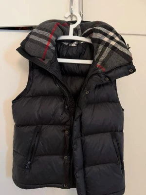 Svart dunväst från burberry - Snygg svart dunväst med dragkedja och knappar framtill. Västen har en avtagbar huva med rutigt foder i grått, rött och vitt. Två dragkedjefickor på sidorna och quiltad design. Perfekt för lager-på-lager och streetstyle.