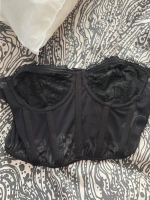 Svart spetskorsett med bygel - Snygg svart korsett med bygel och spetsdetaljer upptill. Modellen är axelbandslös och har förstärkta partier framtill för extra stöd. Materialet är en mix av mesh och spets som ger en lyxig känsla.