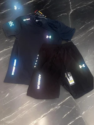 Under Armour i färgen svart reflektiv - Säljer ett svart träningsset från Under Armour med t-shirt och shorts. Båda plaggen har reflekterande loggor och text i regnbågsfärger. Perfekt för gymmet eller löpning. Materialet är lätt och andas, vilket gör det bekvämt under träning. OBS!! Köp inom 48 timmar få 149kr rabatt!!