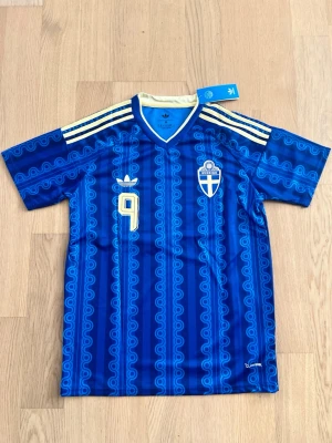Blå Sverige VM 2026 fotbollströja Med Isak  - Retroinspirerad blå fotbollströja från Adidas med Sveriges landslagsemblem, nummer 9 i gult och klassiska Adidas-ränder på axlarna. Tröjan har mönstrad design, V-ringning och är tillverkad i lätt, ventilerande polyester. Perfekt för fotbollsträning eller support.