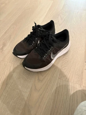 Svarta Nike sneakers med vit sula - Snygga svarta Nike sneakers med vit sula och vit swoosh på sidan. Ovandelen är i mesh och syntetmaterial för bra andningsförmåga. Klassisk låg modell med snörning och bekväm passform, perfekt för en sportig och stilren look.