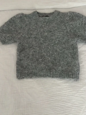 Grå fluffig stickad tröja - Mysig grå stickad tröja med fluffig känsla och rund hals. Tröjan har kortare ärmar och är tillverkad i ett mjukt, luddigt material som ger en avslappnad look. Perfekt för lager-på-lager och chill dagar.