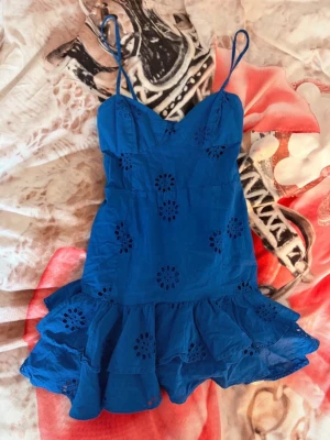 Blå kort klänning från Zara - Supersöt blå klänning från Zara med smala axelband och volangig kjol. Klänningen har broderade blommor och är tillverkad i bomull, perfekt för varma dagar. Snygg v-ringning och luftig passform gör den både bekväm och trendig.