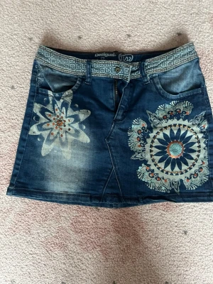 Desiguai jeanskjol med broderi - Cool jeanskjol från Desigual med unika broderade mönster och detaljer framtill. Kjolen har två framfickor, två bakfickor med mönster och ett färgglatt tyg i ena bakfickan. Midjebandet har ett grafiskt tryck och kjolen stängs med dragkedja och knapp.