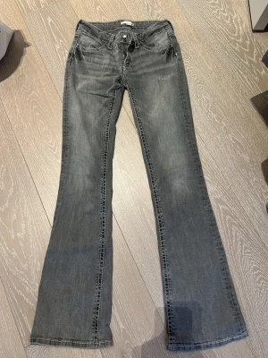 Grå bootcut jeans med kontrastsömmar - Snygga grå bootcut jeans med klassisk femficksdesign och vita kontrastsömmar på bakfickorna. Jeansen har låg midja och lätt slitna detaljer framtill. Perfekta för en avslappnad och trendig look.