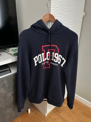 Blå hoodie Polo Ralph Lauren - Mörkblå hoodie från Polo Ralph Lauren med stor vit och röd logga framtill där det står 'POLO 1967'. Huva med dragsko och insida i randigt tyg. Klassisk passform och mjukt material, perfekt för en avslappnad stil.