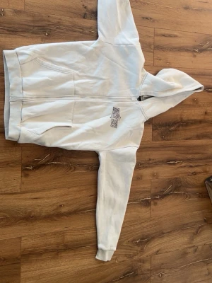 Vit hoodie från SMOG med dragkedja - Säljer en vit hoodie från SMOG i storlek S. Hoodien har dragkedja framtill, två fickor och en broderad logga på bröstet. Tillverkad i mjukt material med borstad insida för extra komfort. Perfekt för en avslappnad och chill stil. Pris kan diskuteras!