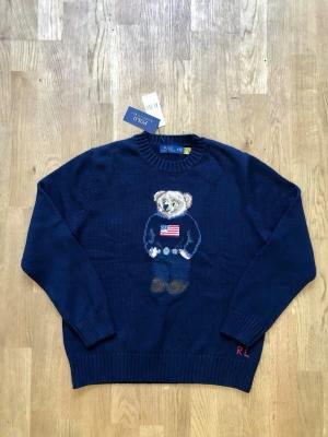 Mörkblå Polo Ralph Lauren björntröja - Stickad mörkblå tröja från Polo Ralph Lauren med ikonisk björn på bröstet, klädd i jeans och tröja med amerikansk flagga. Rund halsringning och ribbade muddar. Broderade initialer 'RL' vid nederkanten. Perfekt för dig som gillar klassisk preppy-stil. Axelbred: 42cm Längd: 63cm