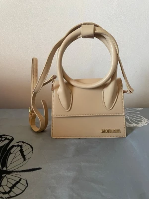 Beige Jacquemus Le Chiquito väska - Supersnygg beige handväska från Jacquemus, modellen Le Chiquito. Väskan har en unik rundad topphandtag, justerbar axelrem och guldfärgad logga framtill. Tillverkad i slätt konstläder med stilren och modern design. Perfekt för dig som vill ha en trendig och ikonisk accessoar.