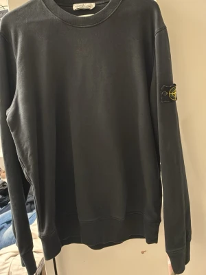 Svart sweatshirt från Stone Island - Svart långärmad sweatshirt från Stone Island med klassisk rund halsringning och ikonisk logotyp-patch på vänster ärm. Tröjan har ribbade muddar vid ärmslut och nederkant. Perfekt för en stilren och avslappnad look.