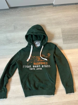 Mörkgrön hoodie Superdry - Mörkgrön hoodie från Superdry med tryck i vitt och orange på bröstet samt texten 'SWEAT. SHIRT. STORE. TOKYO, JAPAN.'. Klassisk känguruficka framtill, vita dragsnören i huvan och ribbade muddar. Perfekt för en avslappnad streetwear-look. Storlek S men passar M utmärkt också 