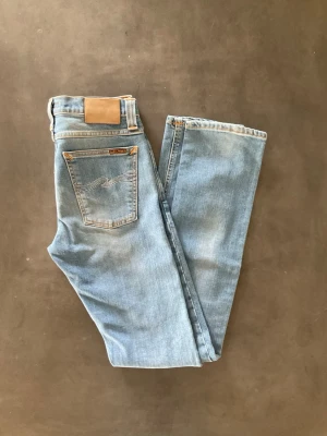 Ljusblå jeans från Nudie Jeans - Säljer ett par ljusblå jeans från Nudie Jeans med klassisk femficksdesign och snygga broderade detaljer på bakfickan. Jeansen har rak passform och är tillverkade i denim av bomull. Perfekta för en avslappnad och trendig look.