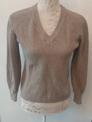 Beige ribbstickad tröja med v-ringning - En stilren beige ribbstickad tröja med v-ringad hals och långa ärmar. Tröjan har breda muddar vid ärmslut och nederkant, vilket ger en snygg passform. 
