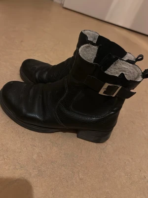 Svarta boots med spänne - Snygga svarta boots i läder med grov sula och silverfärgat spänne på sidan. Dragkedja på insidan för enkel påtagning och en liten ögla bak. Perfekta för dig som gillar en tuffare stil.