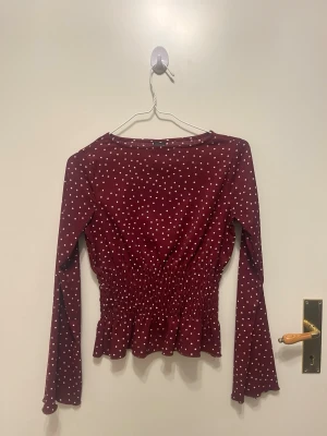 Vinröd prickig blus med smock - Vinröd blus med vita prickar och smockad midja. Långa ärmar och lätt volang nertill ger en söt och trendig look. Perfekt att styla med jeans eller kjol för en cool vardagsoutfit.