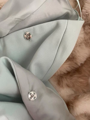 Ljusblå klänning med knappar - Snygg ljusblå klänning med dekorativa knappar framtill och två fickdetaljer. Klänningen har en elegant krage och är tillverkad i ett slätt tyg som ger en stilren look. Perfekt för dig som vill ha en chic och modern stil. Helt ny och oanvänd!🥰