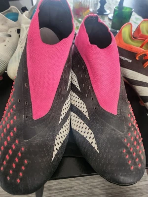 Adidas Predator fotbollsskor svart/rosa - Säljer ett par Adidas Predator fotbollsskor i svart med rosa detaljer och vita ränder på sidorna. Skorna har en strumpliknande ovandel i rosa textil och gummidetaljer för extra grepp på bollen. Sulan har fasta dobbar i svart och rosa för spel på gräs.