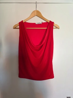 Röd draperad linnetopp från NA-KD - Snygg röd linnetopp från NA-KD med draperad halsringning. Toppen är ärmlös och har en mjuk, följsam passform som passar perfekt till jeans eller kjol. Tillverkad i ett lätt och bekvämt material.