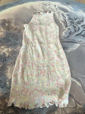 Blommig kort klänning med smock - Supersöt kort klänning med smock och blommigt mönster i pastellfärger. Klänningen har tunna axelband som knyts i nacken och volangkant nedtill. Perfekt för sommarens alla tillfällen!