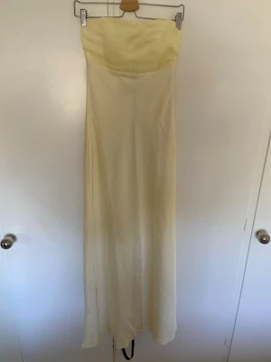 Ljusgul axelbandslös långklänning  - Elegant ljusgul långklänning med axelbandslös design och lätt draperad överdel. Klänningen har en mjuk, flytande siluett. Ljusgult band ingår (se bild)Helt ny. Storlek 34. 