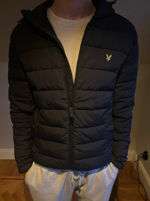 Svart pufferjacka från Lyle & Scott - Svart pufferjacka från Lyle & Scott med guldfärgad logga på bröstet. Jackan har huva, dragkedja framtill och quiltad design. Perfekt för kalla dagar och riktigt skön att bära. Klassisk och stilren look som funkar till det mesta.