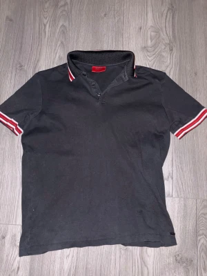 Hugo Boss pikétröja  - Svart pikétröja från Hugo Boss med klassisk krage och knäppning framtill. Kragen och ärmsluten har snygga röda och vita ränder som ger en sportig vibe. Perfekt till jeans eller shorts för en chill look.
