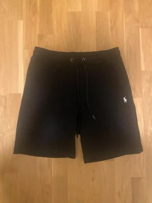 Svarta shorts från Polo Ralph Lauren - Svarta shorts från Polo Ralph Lauren med klassisk logga broderad i vitt på ena benet. Modellen har snörning i midjan och är tillverkad i mjukt bomullsmaterial. Perfekta för en avslappnad och stilren look.