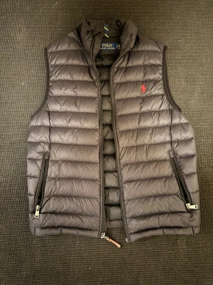 Svart dunväst från Polo Ralph Lauren - Snygg svart dunväst från Polo Ralph Lauren med horisontella sömmar och röd logga på bröstet. Västen har två dragkedjefickor på sidorna och hög krage. Perfekt för lager-på-lager och enkel att matcha med olika outfits. Priset kan diskuteras! Hör av er vid minsta fundering.