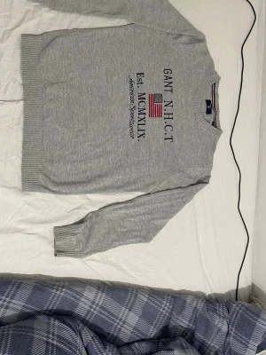 Grå stickad tröja från Gant - Snygg grå stickad tröja från Gant med amerikansk flagga och text på bröstet. Tröjan har rund halsringning, ribbade muddar och nederkant samt en klassisk college-look. Perfekt för dig som gillar stilrena och sportiga plagg.