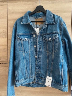 Blå jeansjacka från 157 - Klassisk blå jeansjacka från 157 med raka linjer, silverfärgade knappar och två bröstfickor med lock. Jackan har en tidlös design med krage och synliga sömmar som ger en cool och avslappnad look. Perfekt att slänga över en hoodie eller t-shirt.