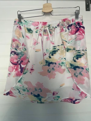 Blommig kortkjol med snörning - Säljer en söt kortkjol med blommigt mönster i rosa, grön, gul och vit. Kjolen har elastisk midja med snörning och är gjord i ett lätt, flowy material som passar perfekt till varma dagar. Omlott-design framtill ger en extra snygg detalj.