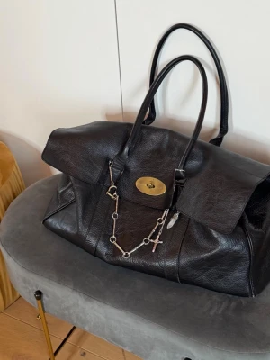 Svart Mulberry Bayswater handväska - Snygg svart Mulberry Bayswater handväska i äkta läder med klassiskt guldigt vridlås och dubbla handtag. Väskan har en kedja med berlocker framtill och en rymlig, strukturerad form. Perfekt för dig som vill ha en ikonisk och stilren väska. Weekend bag, large in size, comes without the chains 