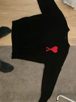 Svart stickad Ami Paris tröja - Svart stickad tröja med ett stort rött hjärta och bokstaven A på bröstet. Tröjan har rund halsringning och långa ärmar. Perfekt för dig som gillar stilrena plagg med en twist. Materialet ser ut att vara mjukt och bekvämt.