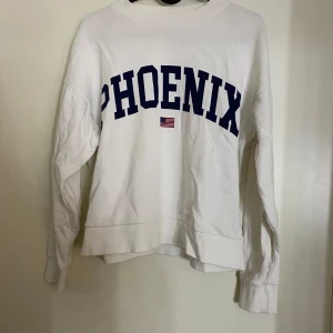 Vit sweatshirt från Gina Tricot - Säljer en vit sweatshirt från Gina Tricot i storlek XXS. Den har texten 'PHOENIX' i mörkblått och en liten amerikansk flagga under. Tröjan är långärmad och perfekt för en avslappnad stil. Superbekväm och lätt att matcha med jeans eller leggings! Knappt använd och är i mycket fint skick. 