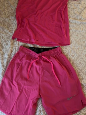 Rosa träningsshorts från Nike - Snygga rosa träningsshorts från Nike med elastisk midja och snörning. Shortsen har en sportig look och är perfekta för aktiva dagar. Tillverkade i ett lätt och snabbtorkande material som passar bra till träning. Tröjan är i storlek L och shortsen i storlek M 