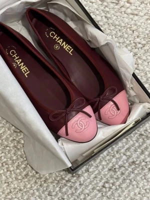 Chanel ballerinaskor vinröd & rosa - Supersnygga ballerinaskor från Chanel i vinrött läder med ljusrosa tåparti och klassisk CC-logga. Skorna har en söt rosett framtill och rundad tå. Perfekta för dig som vill ha en lyxig och trendig touch till din outfit.