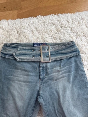 Ljusblå jeans med bredt bälte från secondhand. - Säljer ett par ljusblå jeans från Gang med ett unikt, brett bälte i samma denimtyg och stor silverfärgad spänne. Jeansen har utsvängda ben och klassisk femficksdesign. Perfekta för dig som gillar retrovibbar och vill sticka ut lite extra. Köpt secondhand.