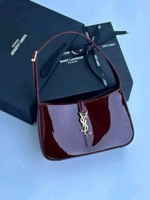 Vinröd Saint Laurent handväska - Snygg vinröd handväska från Saint Laurent i glansigt skinn med guldigt YSL-logo framtill. Väskan har en stilren, rektangulär form och ett kort axelband. Perfekt accessoar för att lyfta din outfit.